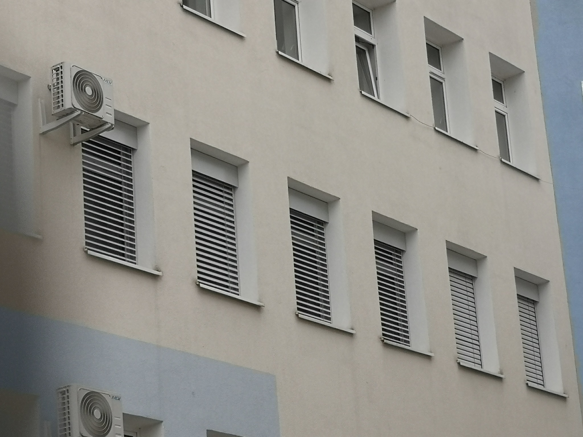 Elewacja budynku z oknami osłoniętymi poziomymi listwami, z zamontowanymi klimatyzatorami na ścianie.