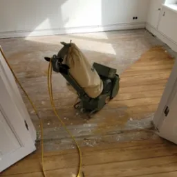 Cyklinowanie podłogi drewnianej w trakcie prac, widoczna maszyna do cyklinowania z workiem na pył i żółtym kablem na tle częściowo wycyklinowanej podłogi i białych ścian.