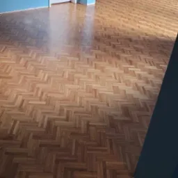 Parkiet ułożony w jodełkę, świeżo po lakierowaniu, z widocznymi refleksami światła, na tle błękitnych ścian i białych drzwi.