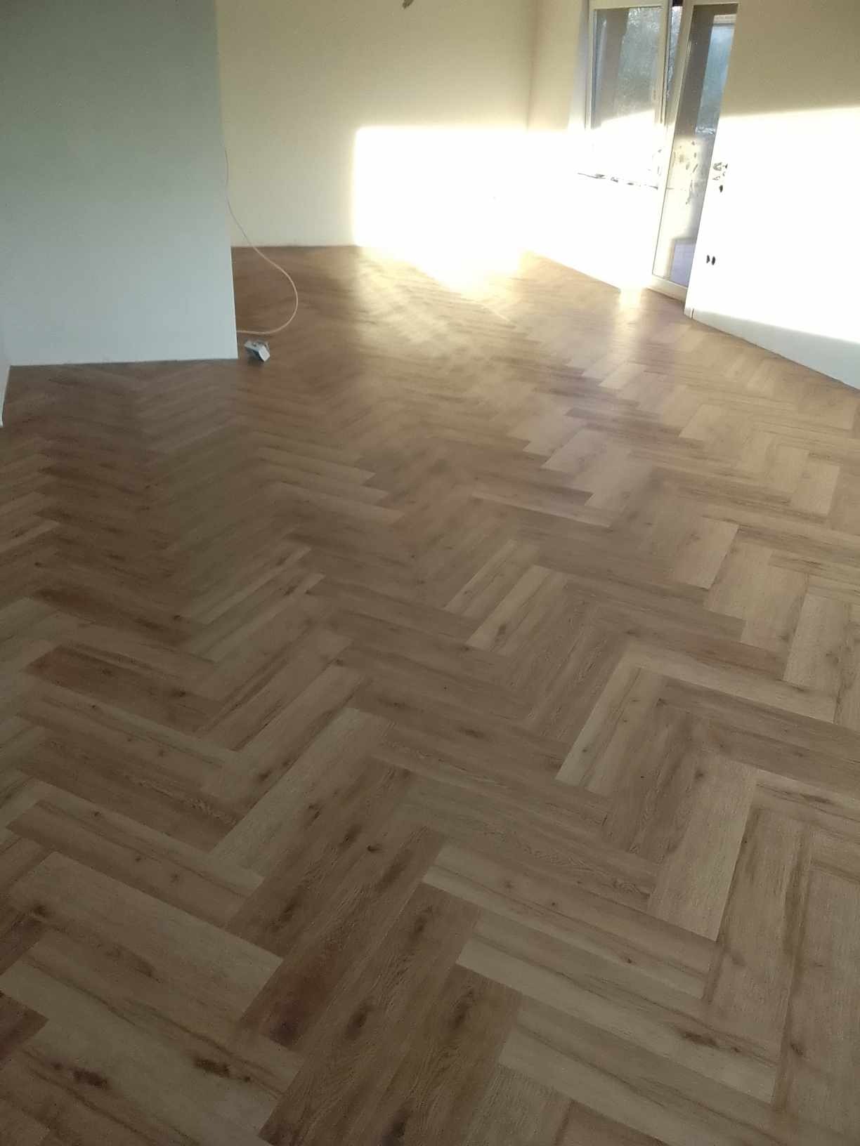 Nowo ułożona podłoga z paneli w jodełkę francuską, widok na jasne, puste wnętrze z otwartymi drzwiami balkonowymi, kabel zwisający ze ściany.