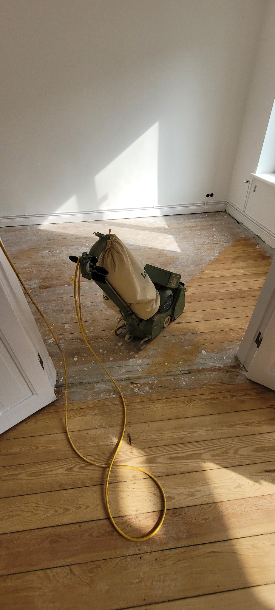 Cyklinowanie podłogi drewnianej w trakcie prac, widoczna maszyna do cyklinowania z workiem na pył i żółtym kablem na tle częściowo wycyklinowanej podłogi i białych ścian.