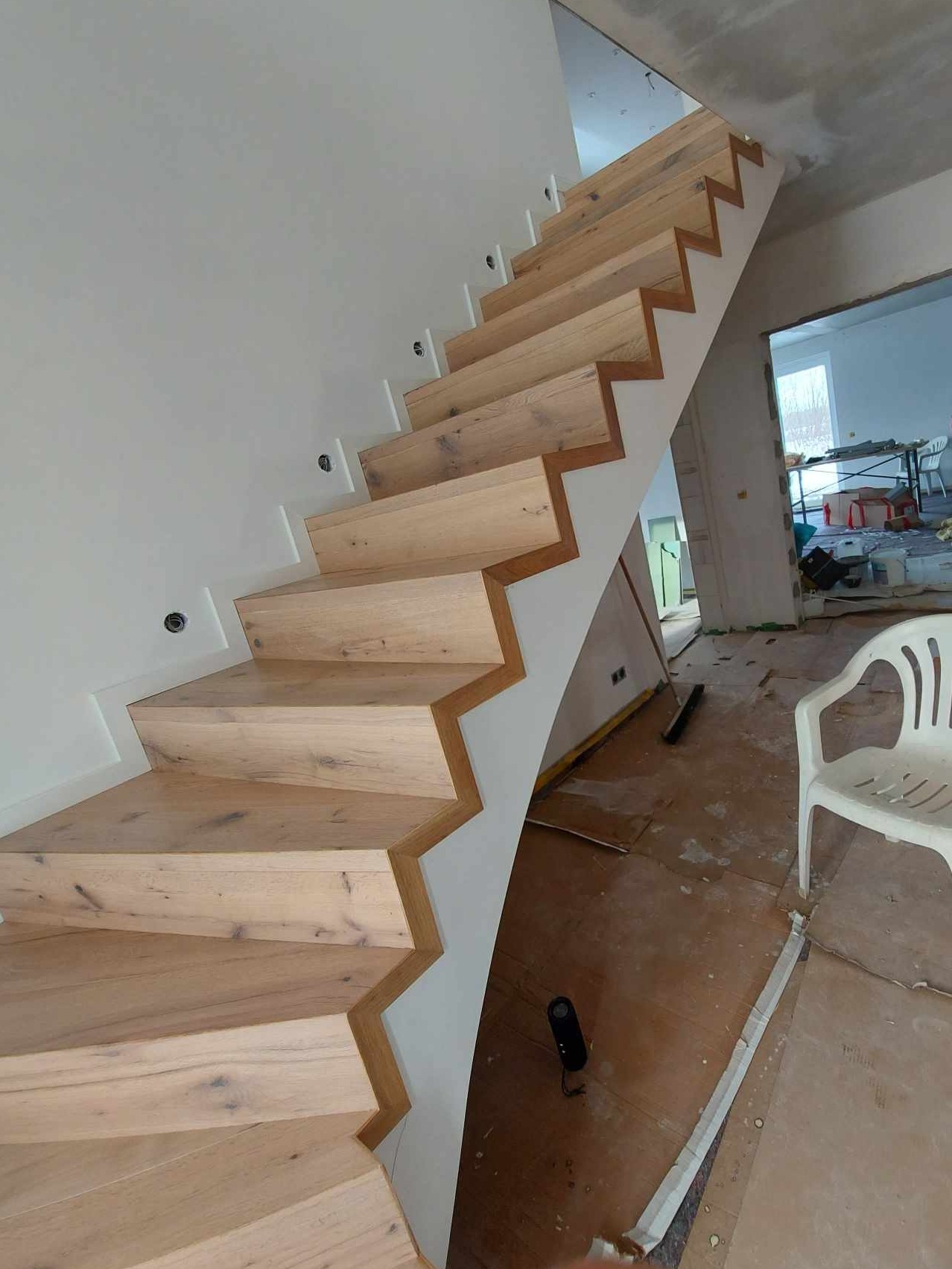 Drewniane schody z jasnego drewna z charakterystycznym zygzakowatym wykończeniem, zamontowane na białej, zakrzywionej konstrukcji. W tle widoczne pomieszczenie w trakcie remontu z niezabezpieczoną...