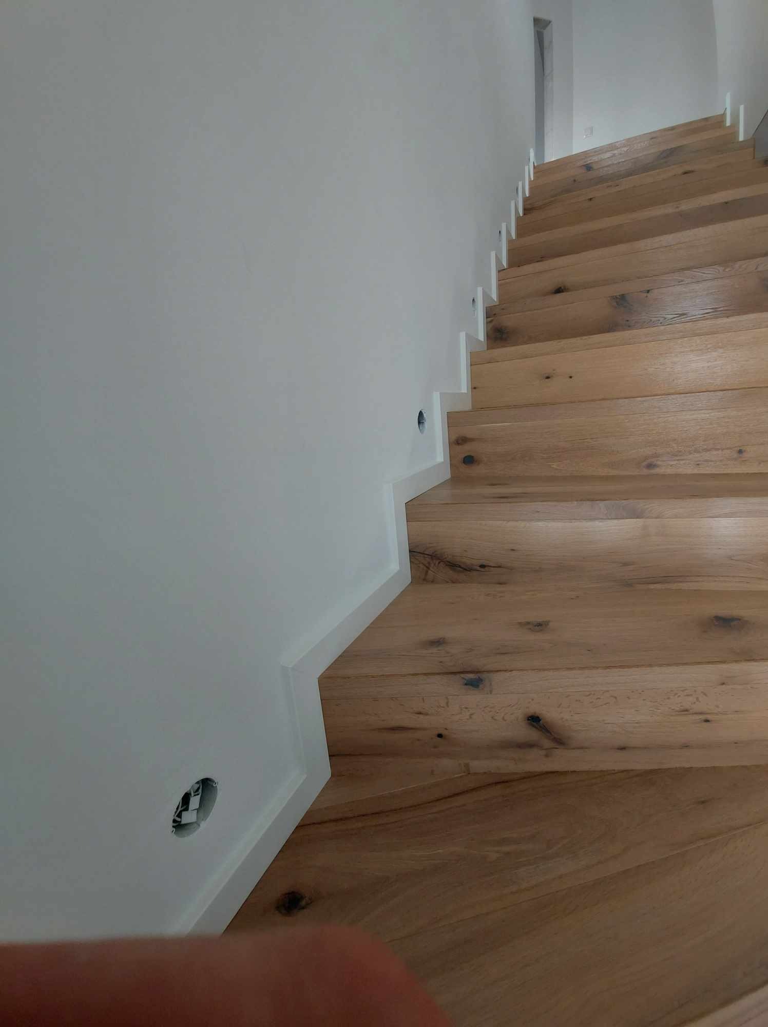 Schody z jasnego drewna dębowego, wykończone białą listwą przypodłogową, z widocznymi otworami na oświetlenie w ścianie.
