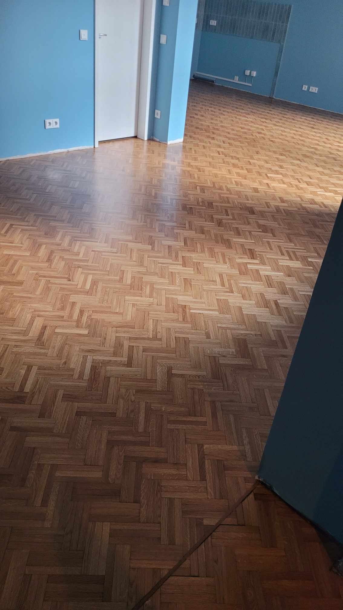 Parkiet ułożony w jodełkę, świeżo po lakierowaniu, z widocznymi refleksami światła, na tle błękitnych ścian i białych drzwi.