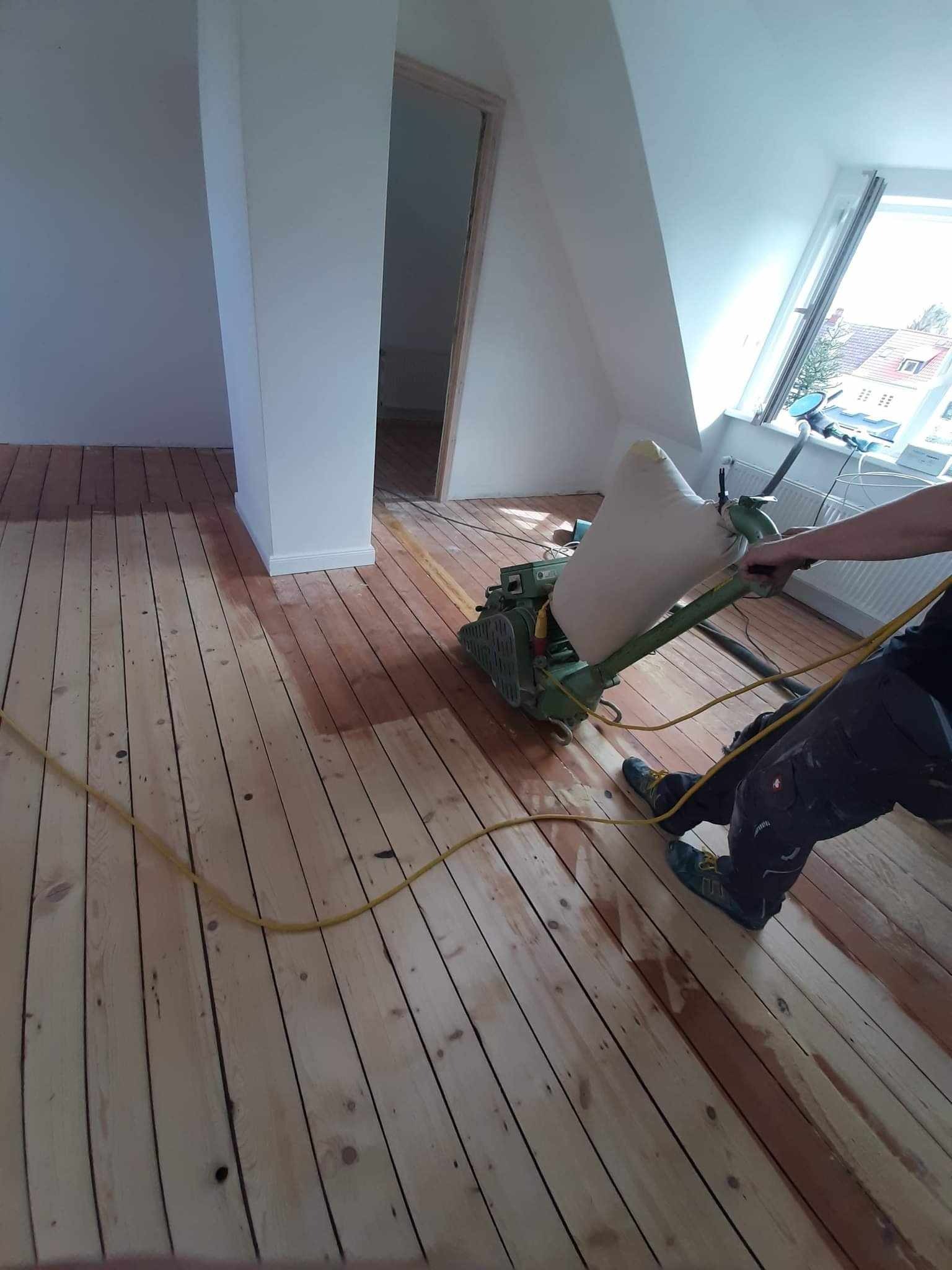 Praca w toku w Łowiczu: parkiet poddawany obróbce szlifierskiej maszyną, widoczne różnice w kolorze drewna przed i po cyklinowaniu. W tle skos dachu i okno.