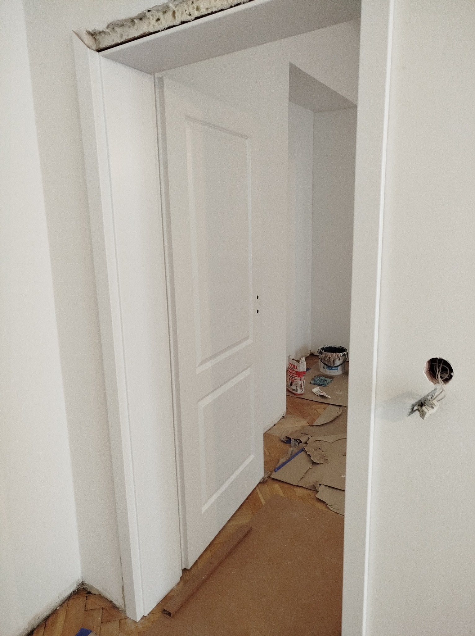 Otwarta, biała, wewnętrzna stolarka drzwiowa z widocznym fragmentem instalacji elektrycznej i zabezpieczonym podłożem w trakcie prac wykończeniowych.