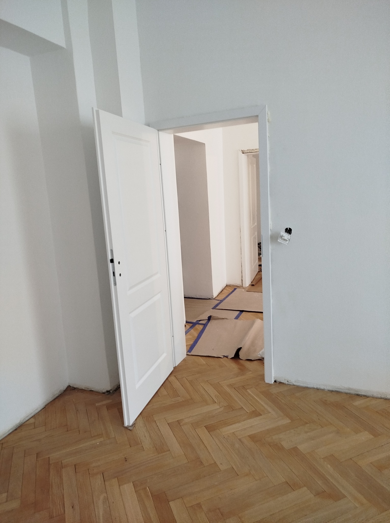 Otwarta biała płycinowa drzwi w odnowionym pokoju z parkietem w jodełkę, zabezpieczonym kartonem i taśmą malarską przed zabrudzeniem podczas prac wykończeniowych, widoczne ślady po gniazdku...