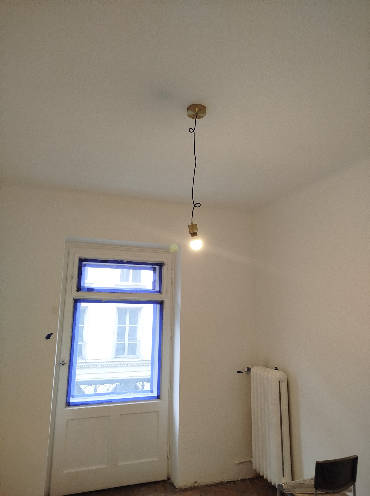 Pokój w trakcie przygotowania do prac wykończeniowych: okno i drzwi zabezpieczone niebieską taśmą malarską, widoczny grzejnik i wisząca żarówka.