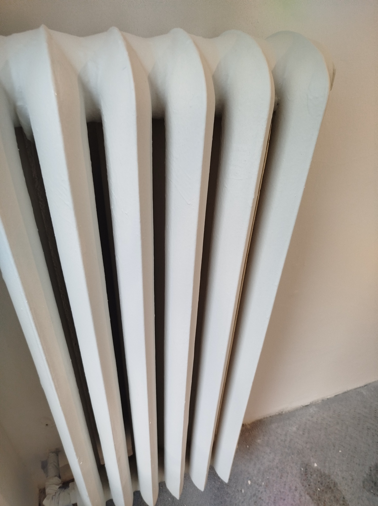 Biały, żeliwny grzejnik żeberkowy po malowaniu, widoczne niedoskonałości na powierzchni farby, ujęcie z dołu, ściana w tle w odcieniu beżu, szara podłoga.