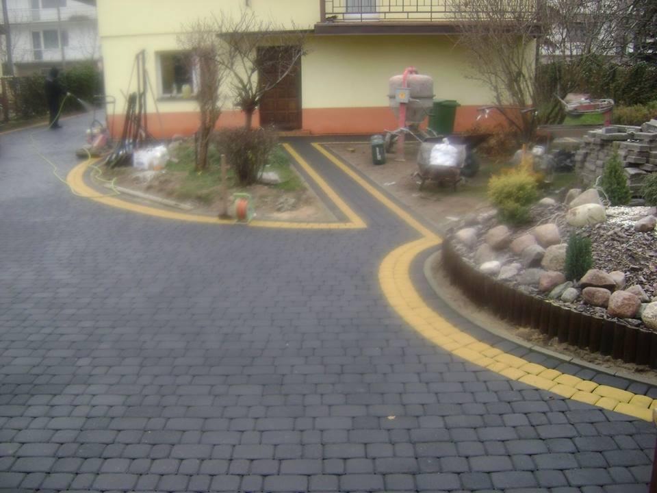 Ułożona ciemnoszara kostka brukowa z żółtymi obrzeżami na podjeździe, w tle dom z żółtą elewacją, częściowo widoczne narzędzia i materiały budowlane, oraz osoba spłukująca powierzchnię wodą.