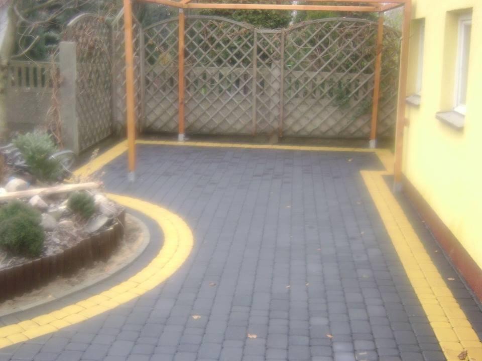 Szara kostka brukowa z żółtymi obramowaniami na podwórku, widoczna pergola z drewnianą kratą w tle, obok żółty budynek z oknem, mały klomb z kamieniami i roślinami.