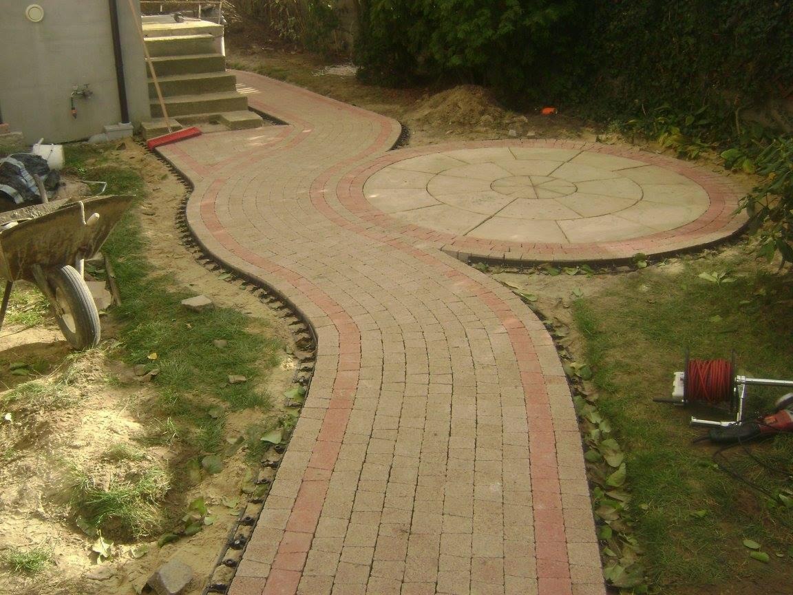 Kręta ścieżka z beżowej kostki brukowej z czerwonym obramowaniem, prowadząca do okrągłego placu z kostki, widok z boku, taczka i narzędzia w tle.