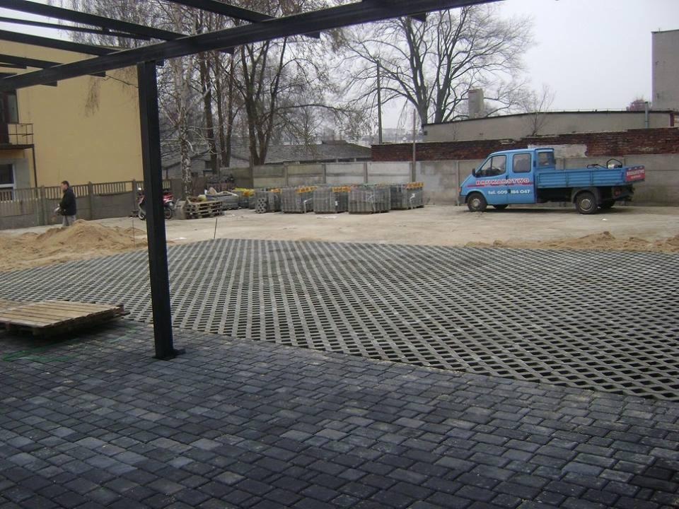 Częściowo ułożona nawierzchnia z ażurowych płyt betonowych i kostki brukowej na placu budowy, z zaparkowanym niebieskim samochodem dostawczym w tle, widok z poziomu gruntu.