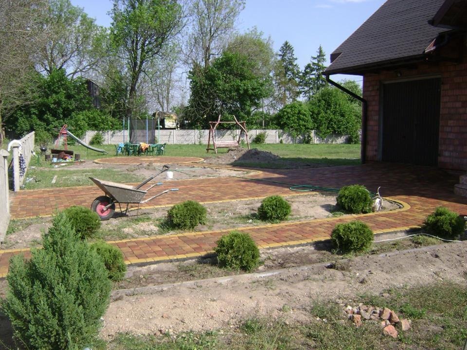 Ułożona ścieżka z czerwono-żółtej kostki prowadząca przez ogród z widoczną taczką, małe krzewy i dom z garażem w tle, w trakcie prac wykończeniowych.