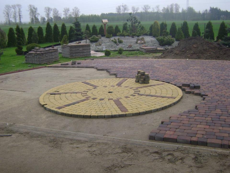 Fragment układanej nawierzchni z kostki brukowej z okrągłym wzorem w centrum, na przygotowanym podłożu z widocznymi zapasami kostki i elementami krajobrazu w tle.