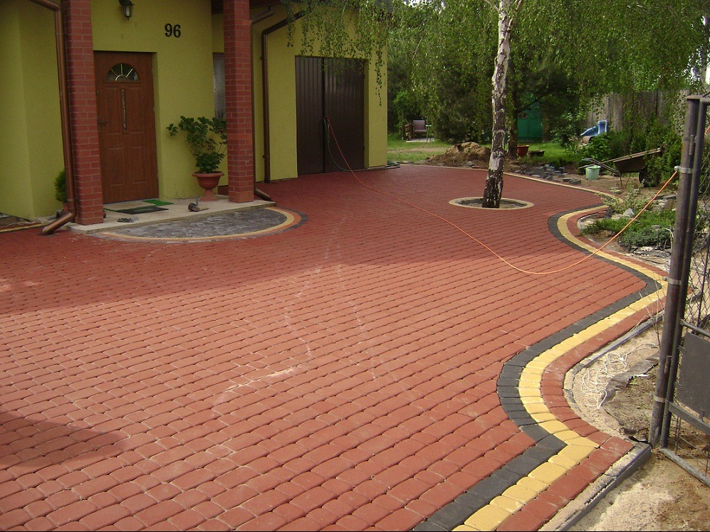 Czerwona kostka brukowa ułożona na podjeździe, z dekoracyjnym paskiem z kostki w kolorze żółtym i czarnym, okalająca rosnące drzewo i prowadząca do domu z numerem 96.