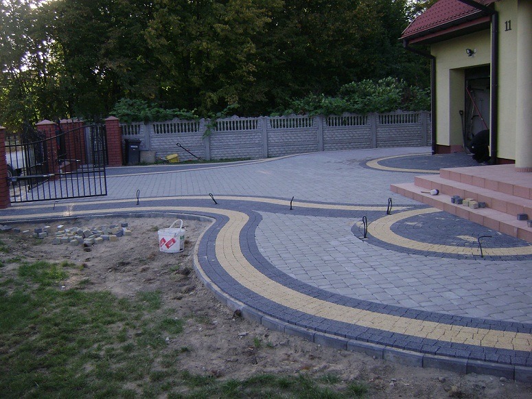 Ułożona z kostki brukowej ścieżka w ogrodzie z dekoracyjnym wzorem z żółtej kostki, widoczne przygotowane kostki do dalszej pracy oraz barierki zabezpieczające.