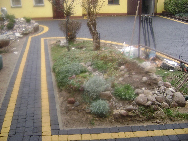 Ułożona czarno-żółta kostka brukowa tworząca ścieżkę obok rabaty z roślinami i kamieniami, widok z góry na posesję z żółtym budynkiem w tle.