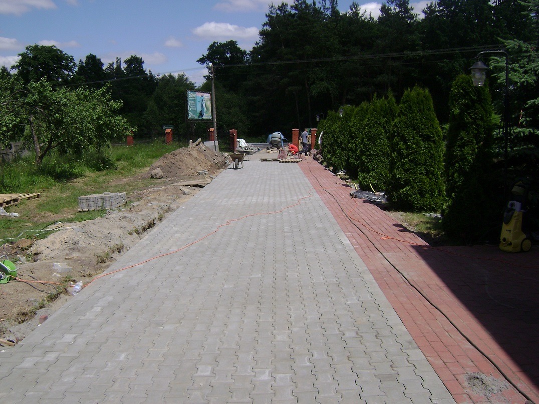 Szeroki podjazd z szarej kostki brukowej w Sieradzu, obrzeża z czerwonej kostki, widoczne prace wykończeniowe i ekipa budowlana w tle, teren jeszcze nie do końca uporządkowany.