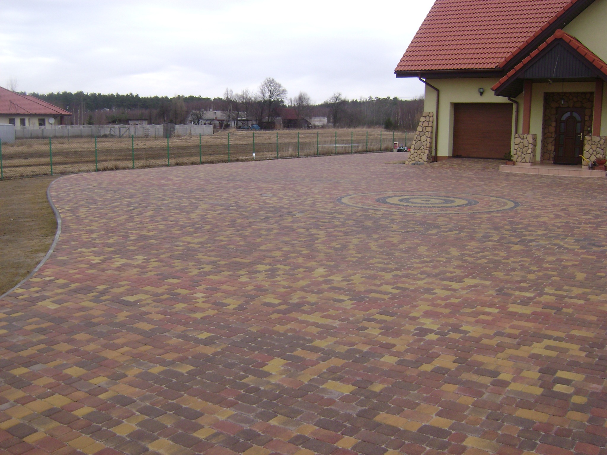 Szeroki podjazd z wielobarwnej kostki brukowej ułożonej w łuk, z dekoracyjnym okręgiem pośrodku, prowadzący do domu z brązową bramą garażową i wejściem obłożonym kamieniem.