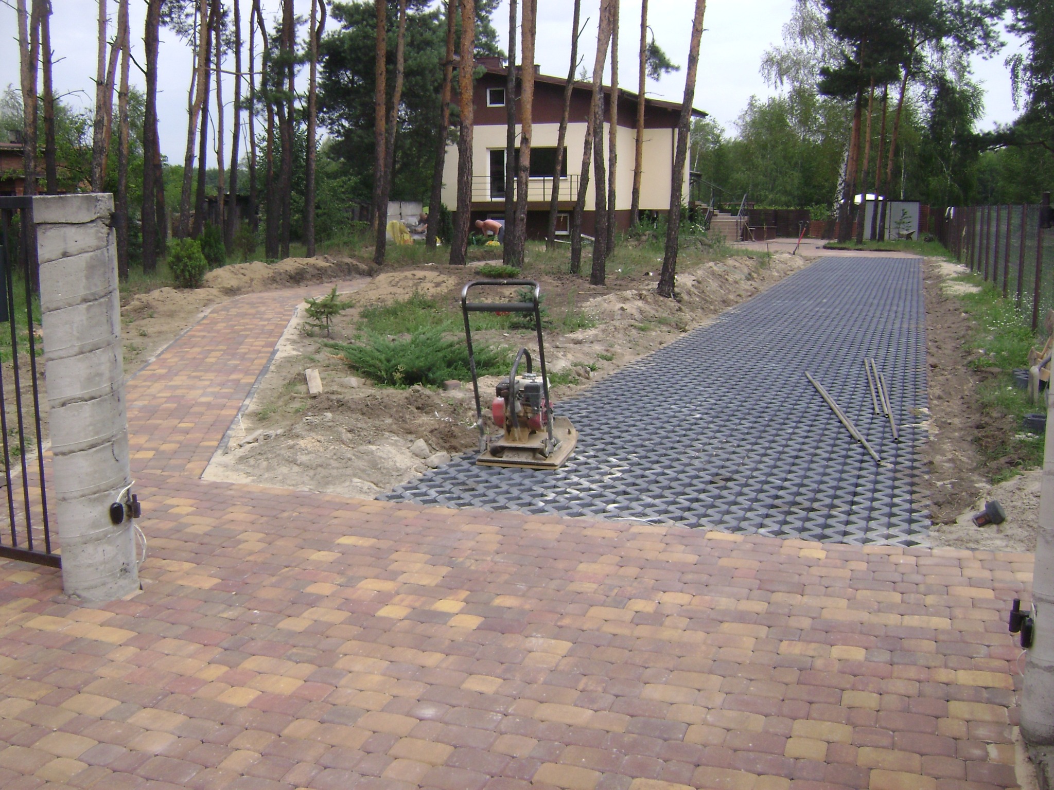 Wyrównywanie terenu pod budowę podjazdu z kostki brukowej, widoczny ubijak mechaniczny, częściowo ułożona kostka ażurowa oraz fragment podjazdu z kolorowej kostki brukowej w odcieniach czerwieni...