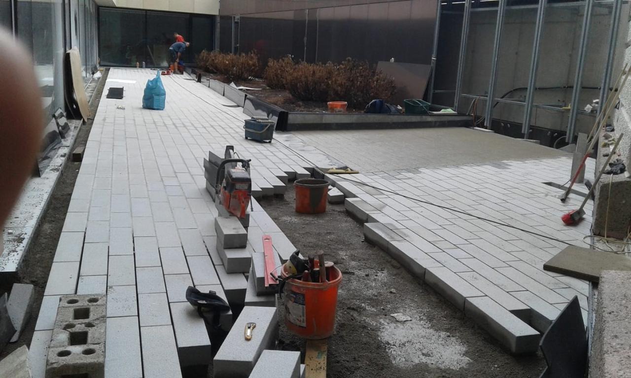 Trwa układanie szarej, prostokątnej kostki na ścieżce; widoczna piła do cięcia kostki, wiadro z narzędziami, częściowo ułożona powierzchnia i pracownik w oddali.