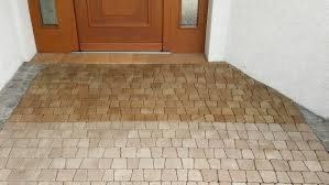 Ułożona kostka w odcieniach brązu i beżu przed wejściem do domu, z widocznym drewnianym progiem drzwi i fragmentem białej ściany. Kostka ułożona w dwa różne wzory, tworząc wyraźne rozgraniczenie...