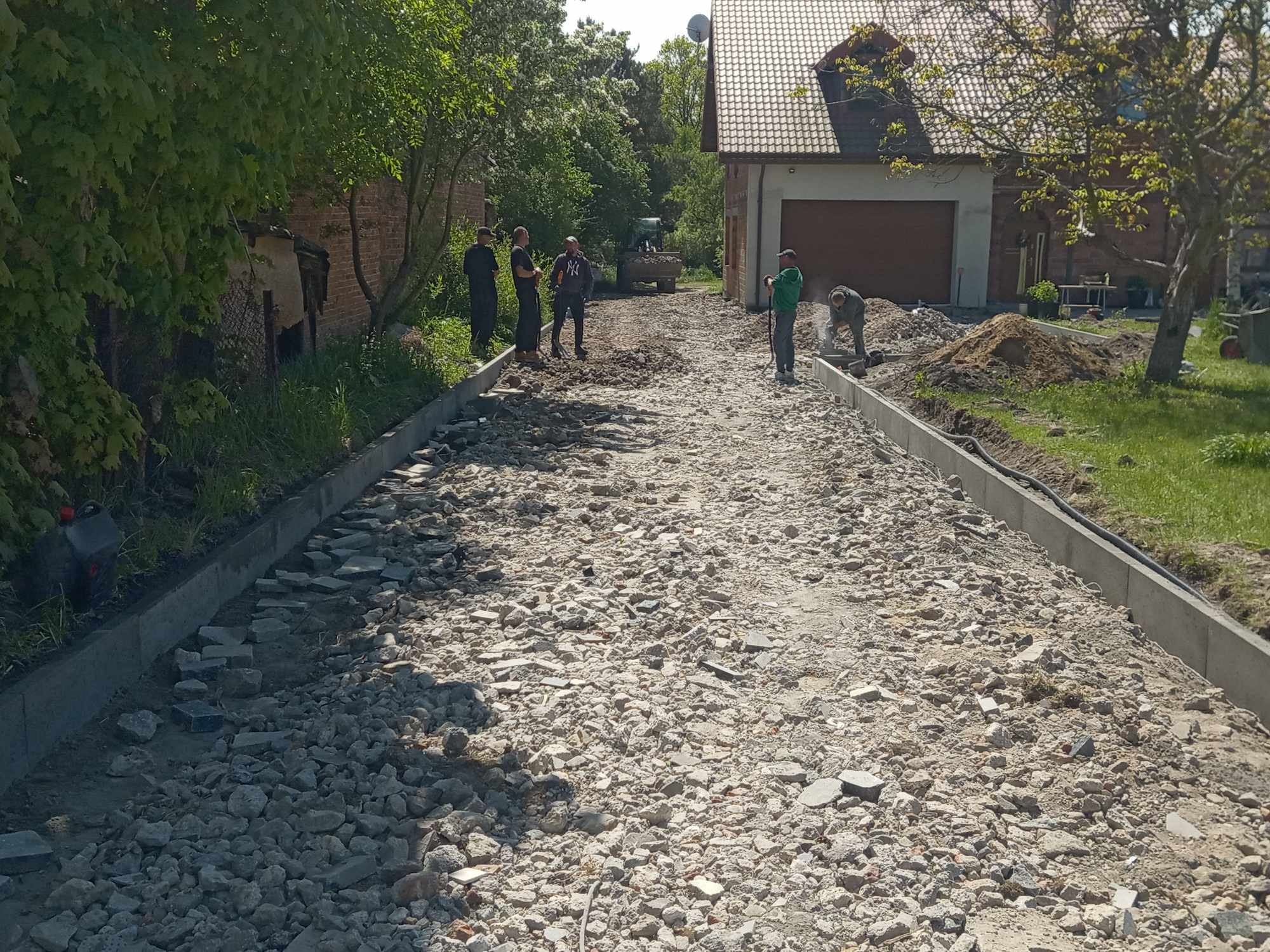 Przygotowanie podłoża pod kostkę brukową z obrzeżami. W tle ekipa i sprzęt budowlany. Widok na posesję z domem i garażem.
