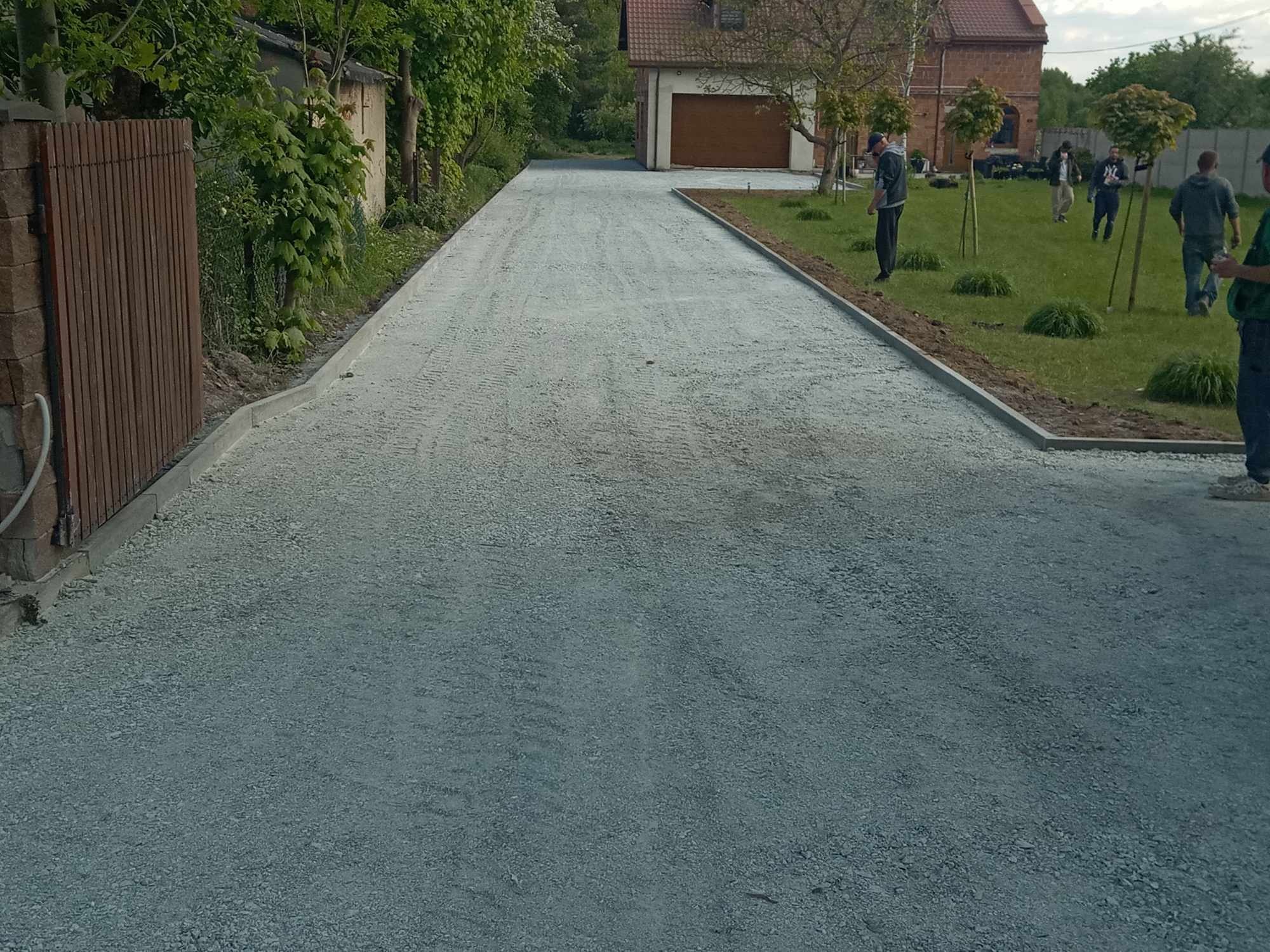 Utwardzona droga dojazdowa z obrzeżami, świeżo po niwelacji, z widocznym budynkiem mieszkalnym w tle i pracownikami na trawniku obok.