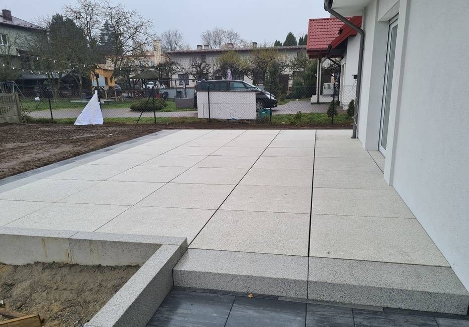 Jasnoszary taras z płyt granitowych, z widocznymi fugami i obrzeżami, przylegający do białego budynku. Częściowo widoczny teren przed posesją.