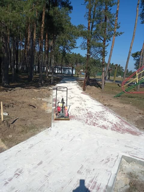 Czerwona kostka brukowa częściowo pokryta białym piaskiem, zagęszczarka w tle, otoczenie leśne i plac zabaw. Widok na ścieżkę w trakcie budowy.