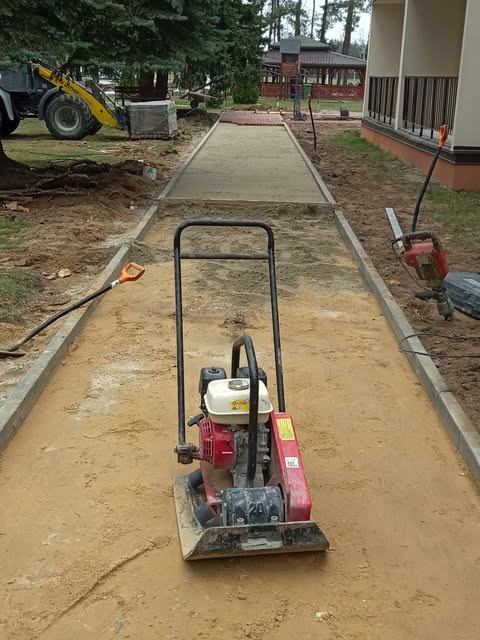 Wyrównywanie podłoża pod ścieżkę z kostki brukowej za pomocą zagęszczarki. W tle widoczny sprzęt budowlany i częściowo ułożona kostka. Prace ziemne w toku.