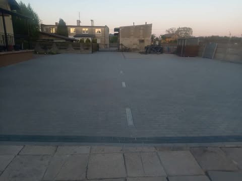 Szara kostka brukowa na posesji z widocznym odpływem liniowym i budynkami w tle, sfotografowana w pochmurny dzień. Widok z poziomu gruntu.