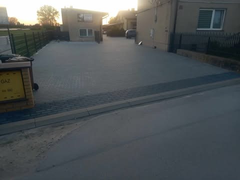 Ułożona kostka brukowa na podjeździe przed budynkiem, widok z poziomu ulicy. Szara kostka, obrzeża z ciemniejszej kostki. Widoczne budynki i ogrodzenie.