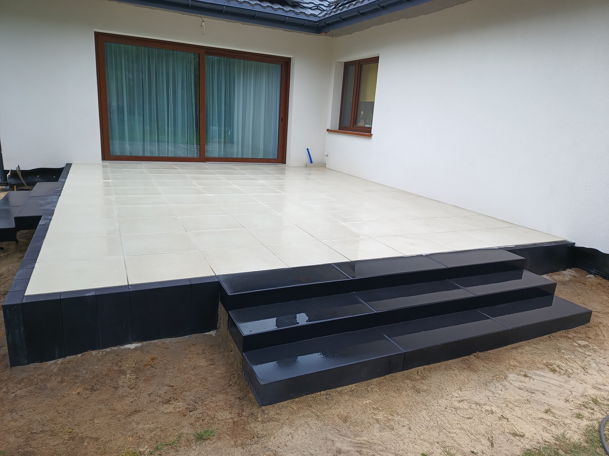 Wykończony taras z jasnych płytek i czarnych schodów, przylegający do białego budynku z oknami. Widoczne detale wykończenia i świeżo ułożona nawierzchnia.