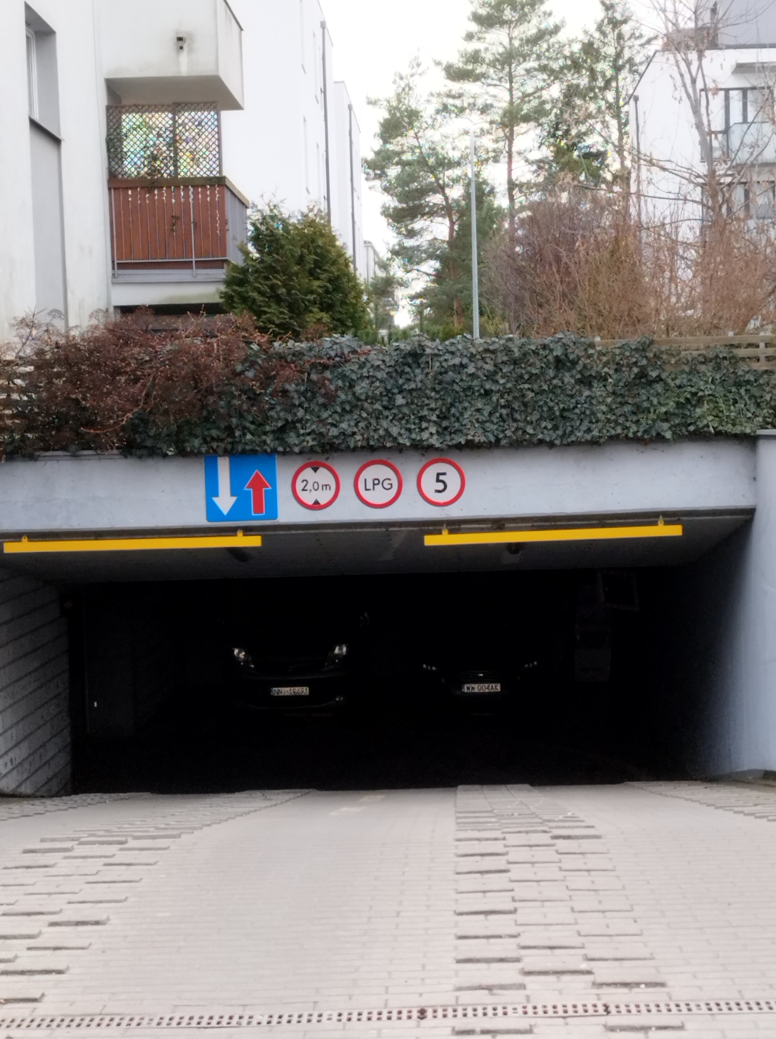 Wjazd do podziemnego parkingu z betonową nawierzchnią, oznakowany ograniczeniem wysokości 2.0m i dopuszczeniem LPG, z zielenią na górze.