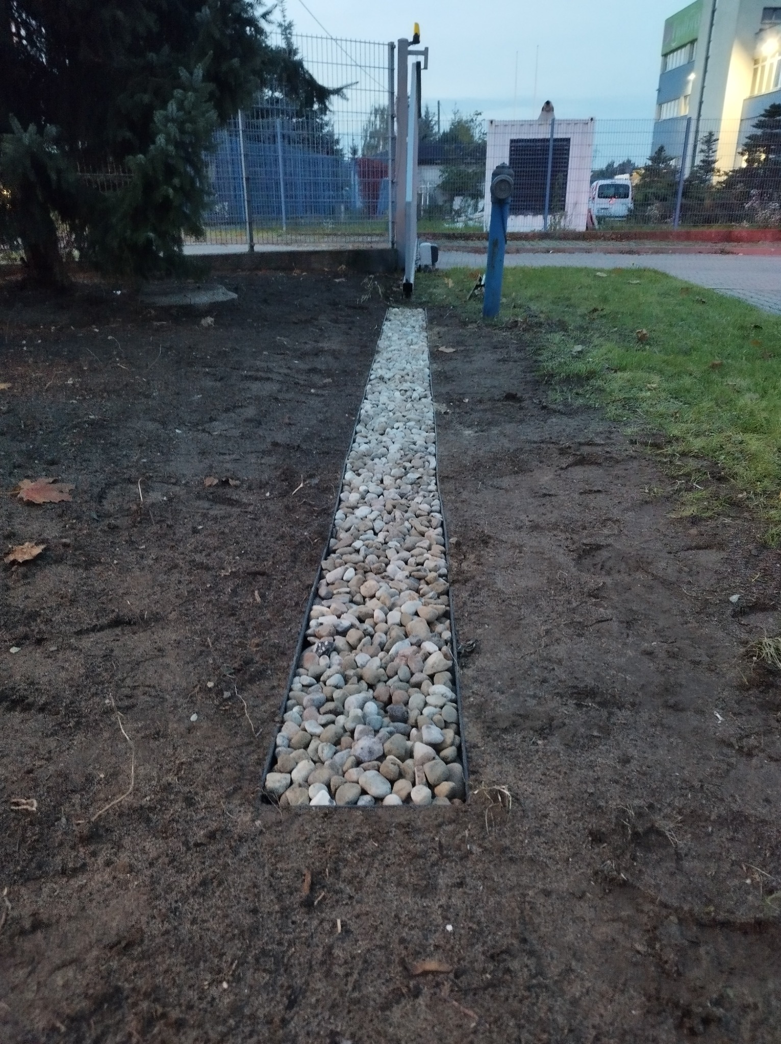 Drenaż liniowy wypełniony jasnymi otoczakami, wkopany w ziemię, z widocznym trawnikiem i budynkiem w tle.