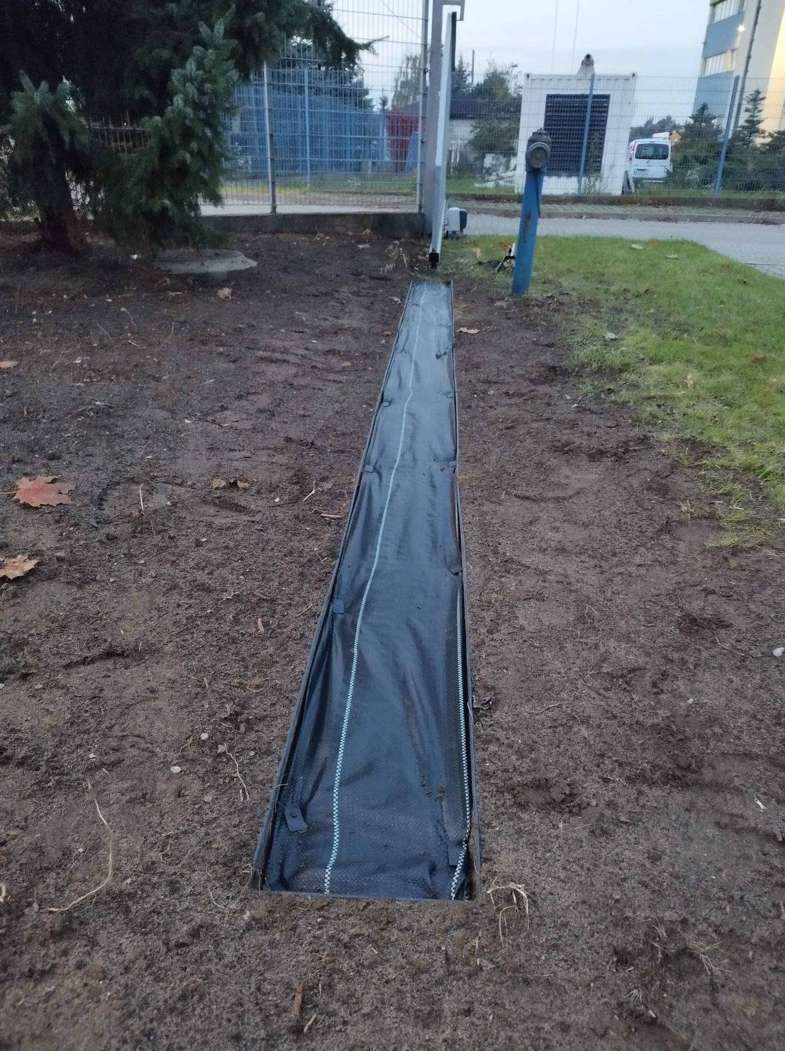 Wykopany rów wyłożony czarną agrowłókniną w ziemi, widok z góry, w tle trawnik i ogrodzenie.