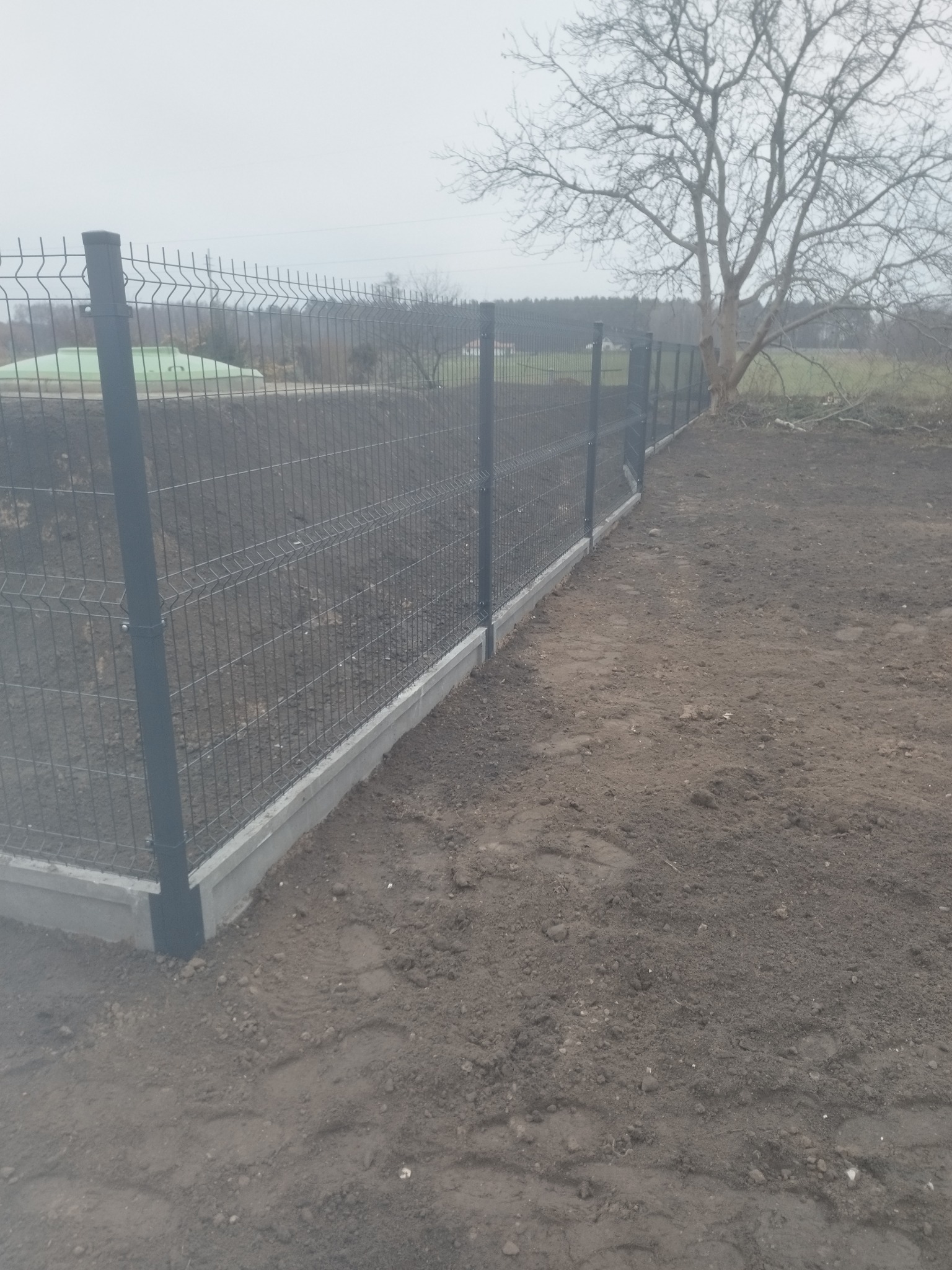 Nowoczesne ogrodzenie panelowe z podmurówką w Błoniu. Metalowa siatka ogrodzeniowa na betonowej podstawie, widok z boku, szary dzień.