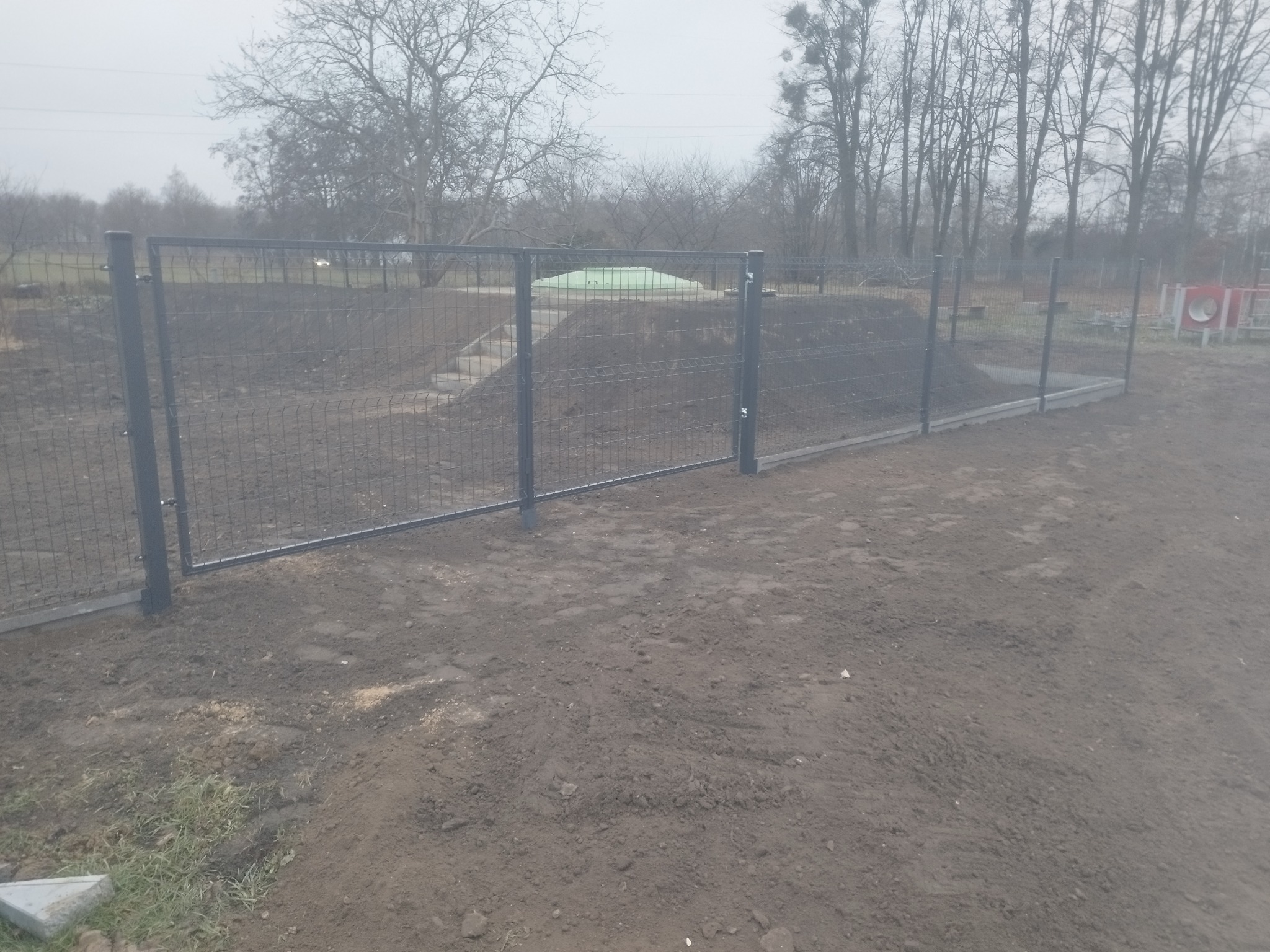 Nowoczesne ogrodzenie panelowe z metalową siatką na betonowej podmurówce, widok na park w tle, pochmurny dzień.