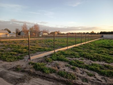 Montaż ogrodzenia z siatki na betonowej podmurówce. Widoczne słupki i częściowo rozciągnięta siatka. Trawa i zabudowania w tle, ciepłe światło zachodzącego słońca.