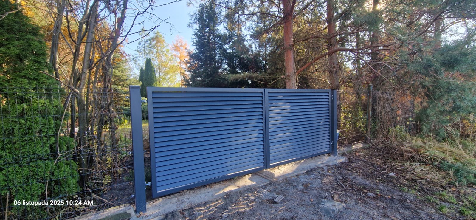 Nowoczesna, przesuwna brama lamelowa w kolorze grafitowym, zamontowana na betonowej podmurówce, otoczona zielenią i drzewami w jesiennej scenerii.