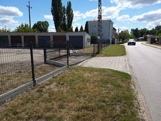 Nowoczesne ogrodzenie z siatki, z betonową podmurówką, oddzielające posesję od drogi. Widoczny fragment trawnika i budynki w tle. Ujęcie z poziomu gruntu.