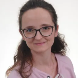 Portret uśmiechniętej kobiety w okularach i różowej bluzce z ozdobnym sznurowaniem, na jasnym tle. Widoczna delikatna biżuteria.