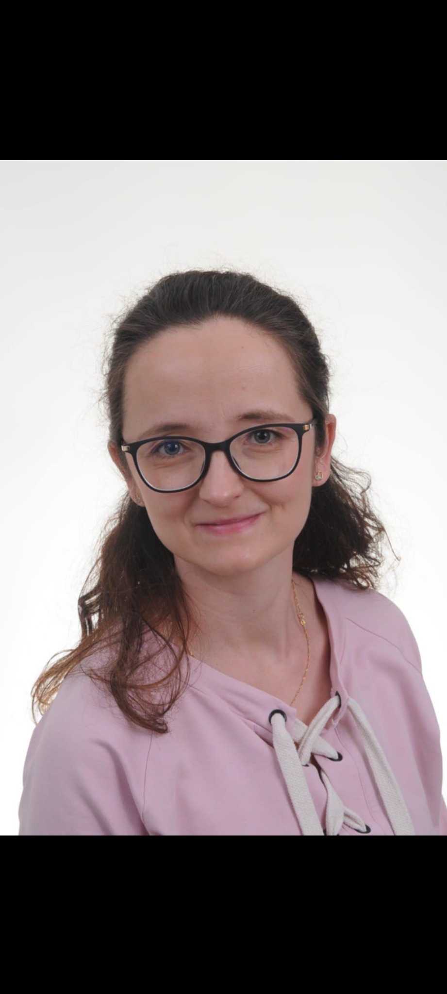 Portret uśmiechniętej kobiety w okularach i różowej bluzce z ozdobnym sznurowaniem, na jasnym tle. Widoczna delikatna biżuteria.
