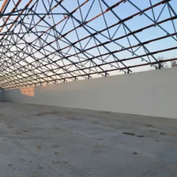 Konstrukcja stalowa dachu w hali produkcyjnej, widok od wewnątrz na szkielet dachu i betonową posadzkę. Widoczny fragment ściany bocznej hali.