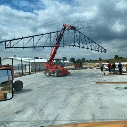 Twój Dach - Montaż stalowej więźby dachowej dźwigiem Maniscopic na placu budowy. Widoczni pracownicy i elementy konstrukcyjne budynku. Ujęcie z kabiny pojazdu, odbicie nieba w szybie.