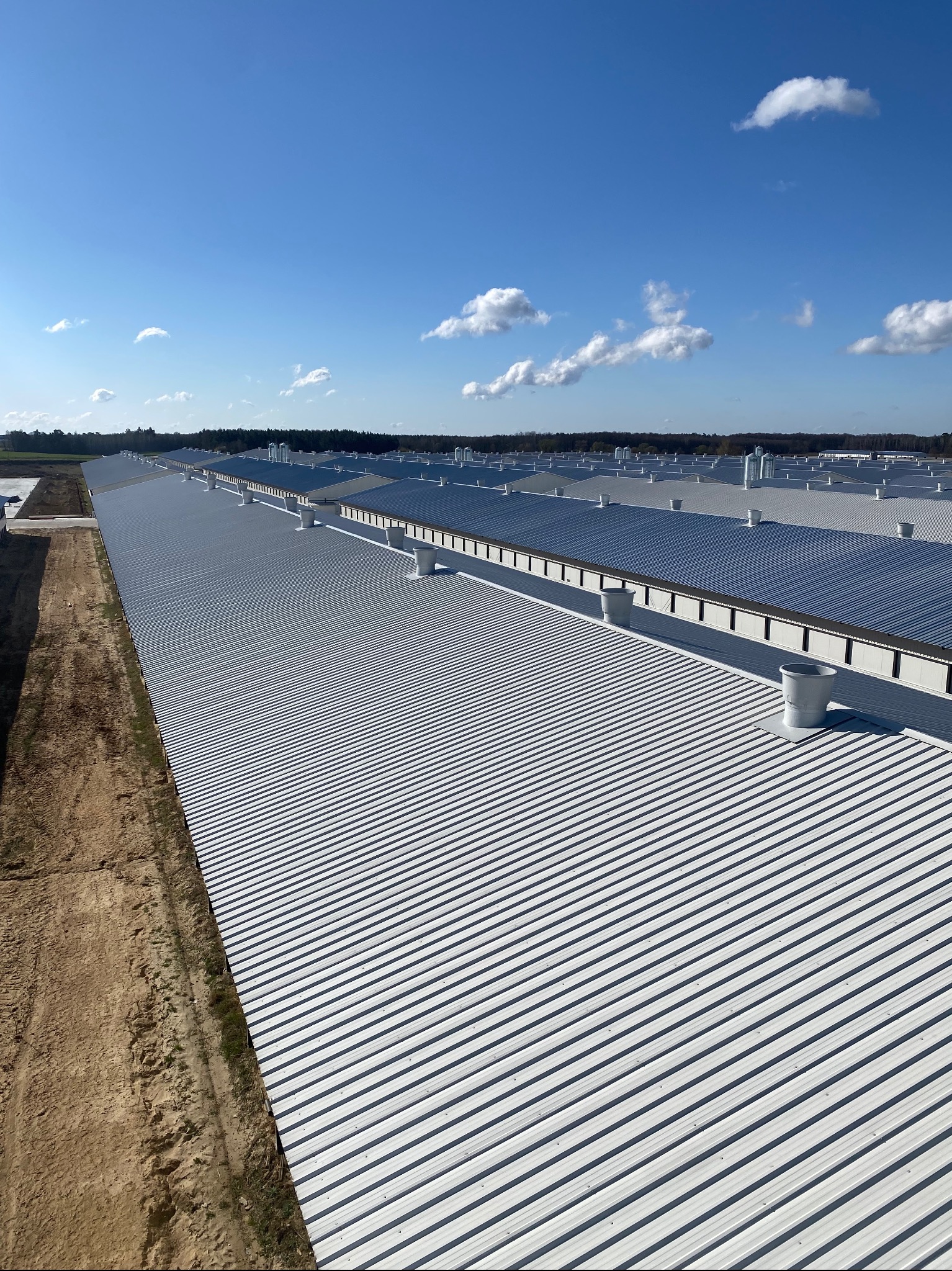Rozległy dach z blachy trapezowej, widok z góry, z licznymi kominami wentylacyjnymi, na tle błękitnego nieba z lekkimi obłokami. Widoczny fragment terenu budowy.