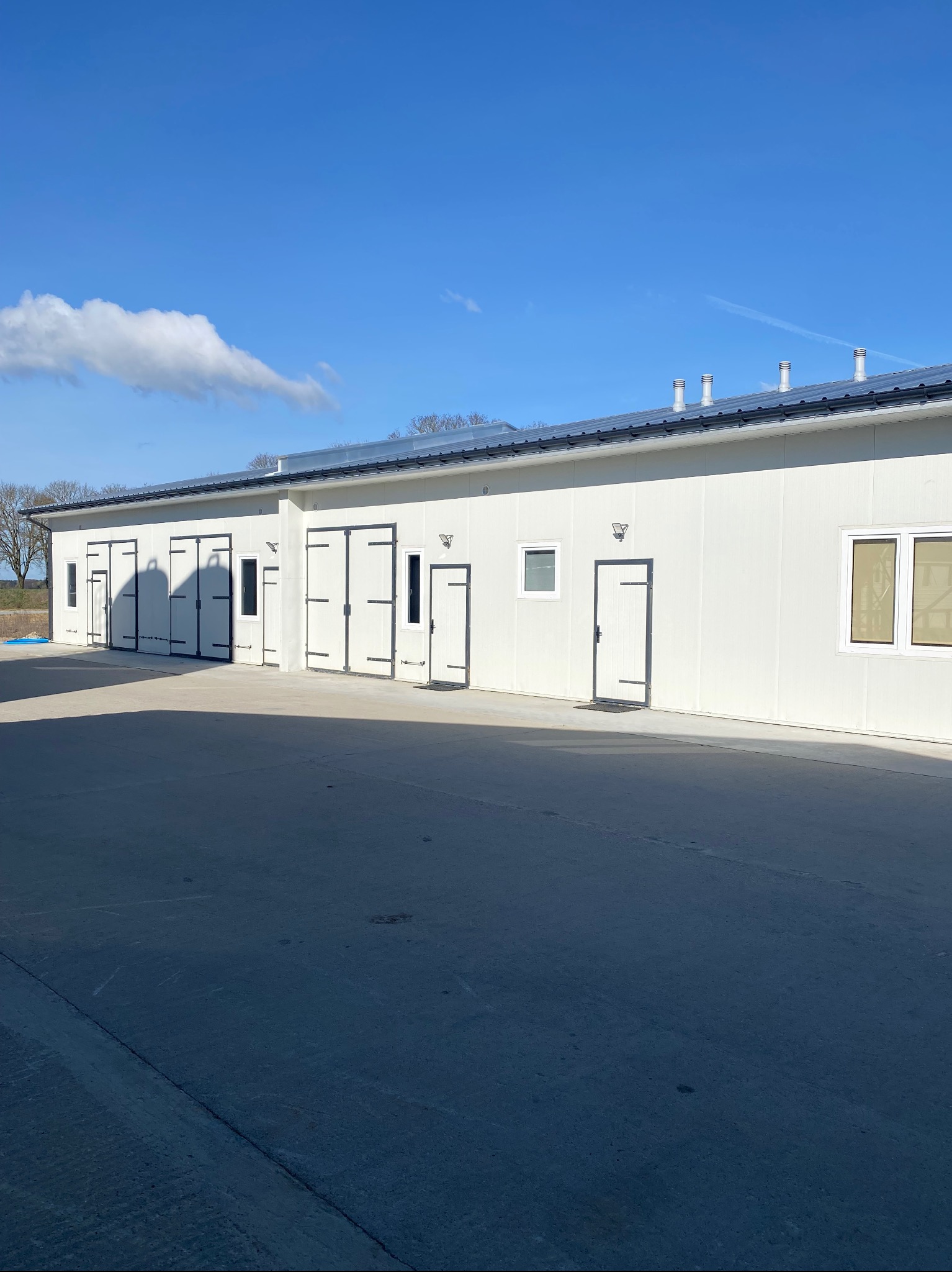 Biały budynek z szarym dachem i otwartymi drzwiami garażowymi. Widoczne kominy na dachu i fragment niebieskiego nieba z chmurami. Budynek w stylu przemysłowym.