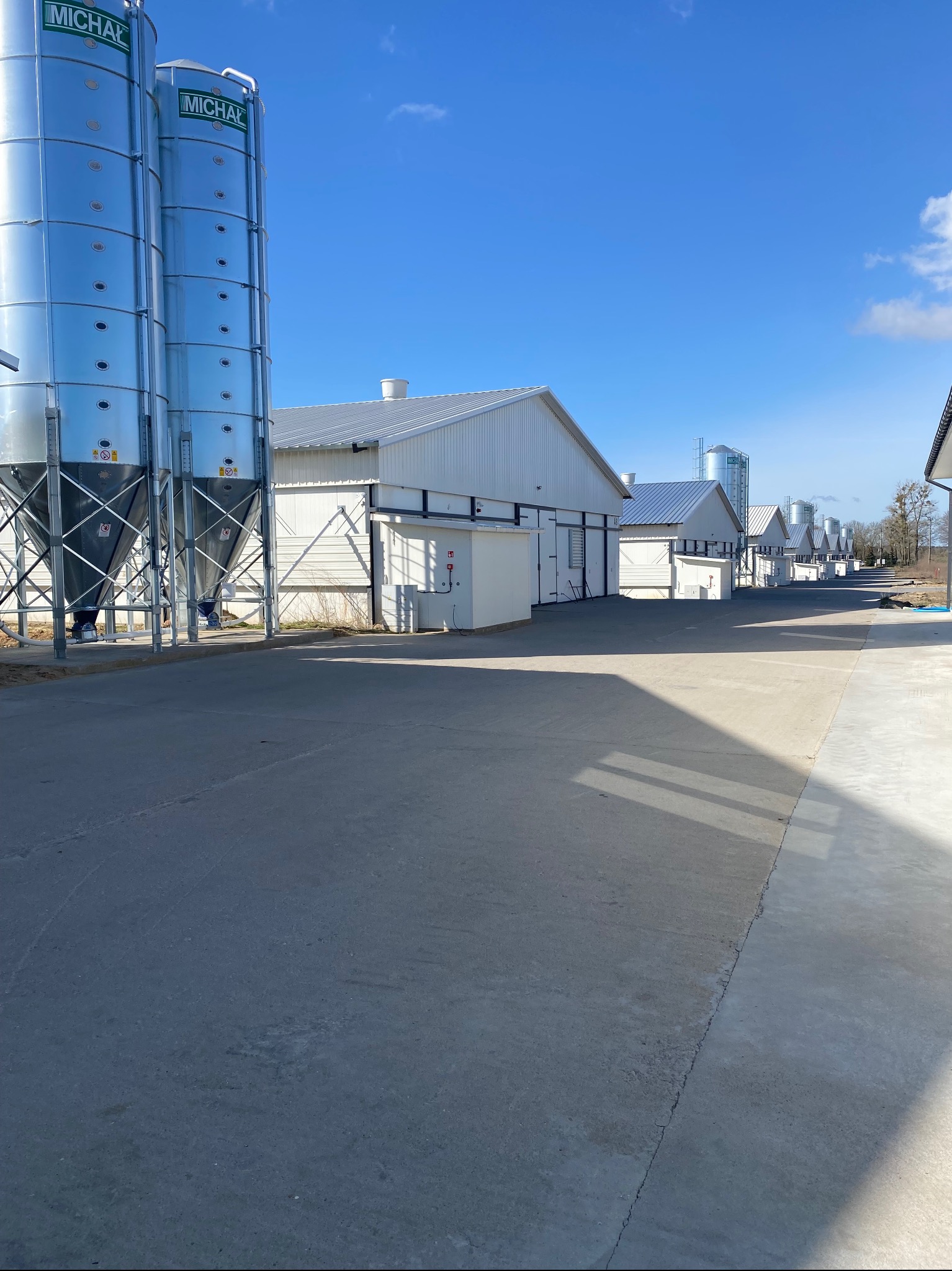 Długi rząd białych budynków z blachy falistej pod błękitnym niebem, z dwoma srebrnymi silosami po lewej stronie. Dachy budynków są również z blachy falistej.